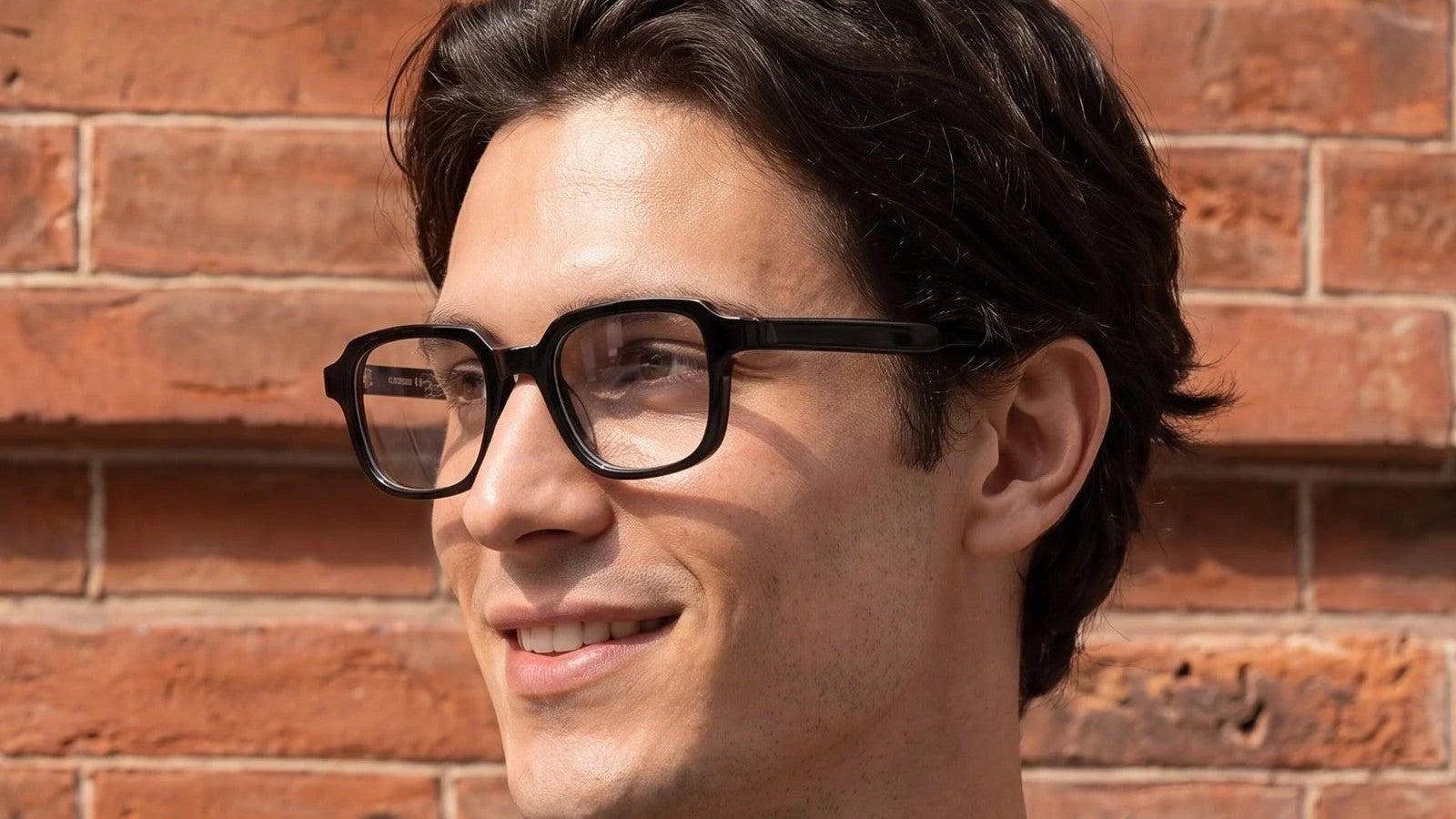 Man in black acetate rectangle glasses smiling in front of a brick wall