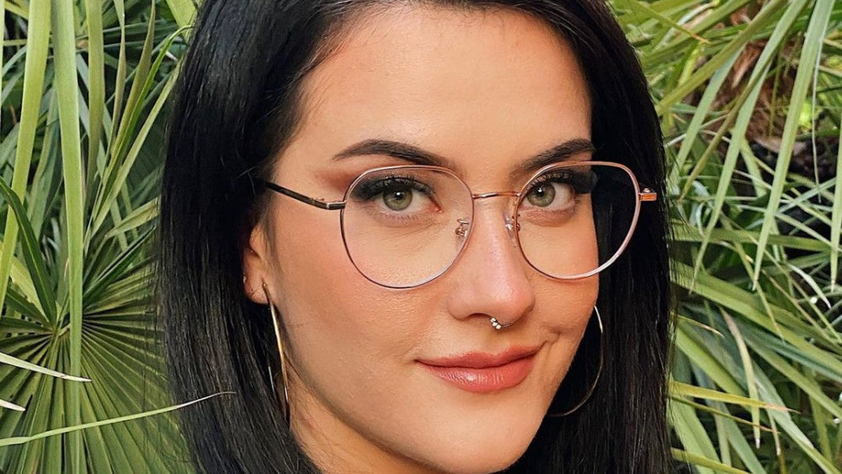 Woman wearing thin rose gold geometric eyeglasses in front of green palm leaves