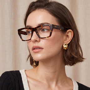 A close-up of a woman wearing rectangular tortoiseshell eyeglasses with the brand name IGIOO visible on the temple arm, styled with gold earrings against a neutral beige curtain background.