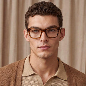 A close-up portrait of a man wearing thick, square tortoiseshell acetate eyeglasses with a warm brown and amber tone men brown eyeglasses