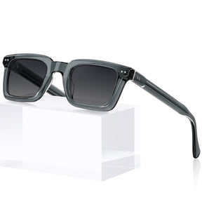 Gray acetate square sunglasses with dark lenses, displayed on a white geometric block against 