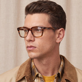 A man wearing tortoiseshell acetate eyeglasses with a subtle 'CATO' brand logo on the temple, styled with a brown corduroy jacket and a yellow shirt men model