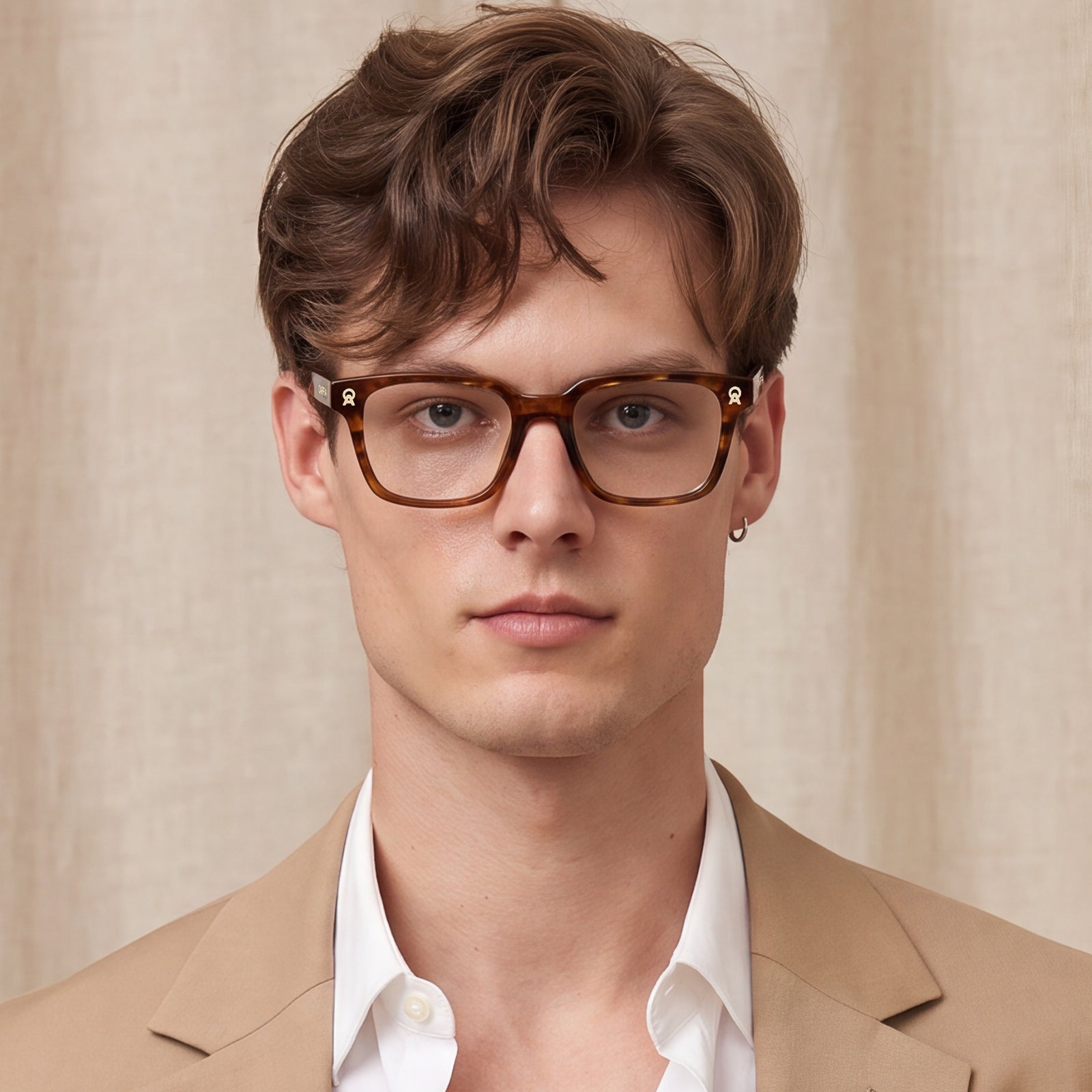 A man wearing brown tortoiseshell square eyeglasses, styled with a beige blazer and white collared shirt against a neutral beige curtain background.