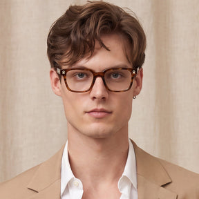 A man wearing brown tortoiseshell square eyeglasses, styled with a beige blazer and white collared shirt against a neutral beige curtain background.